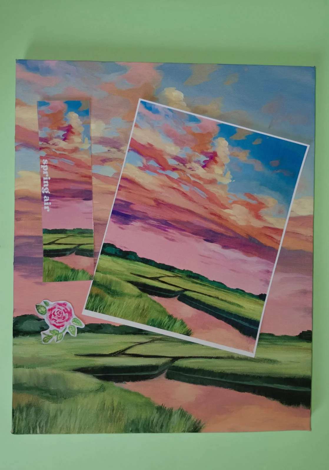 Pintura em tela de um pôr do sol rosa e laranja sobre um campo verde cortado por um rio, com o print e o marcador "spring air" colados por cima e um adesivo de rosa, sobre fundo verde menta.