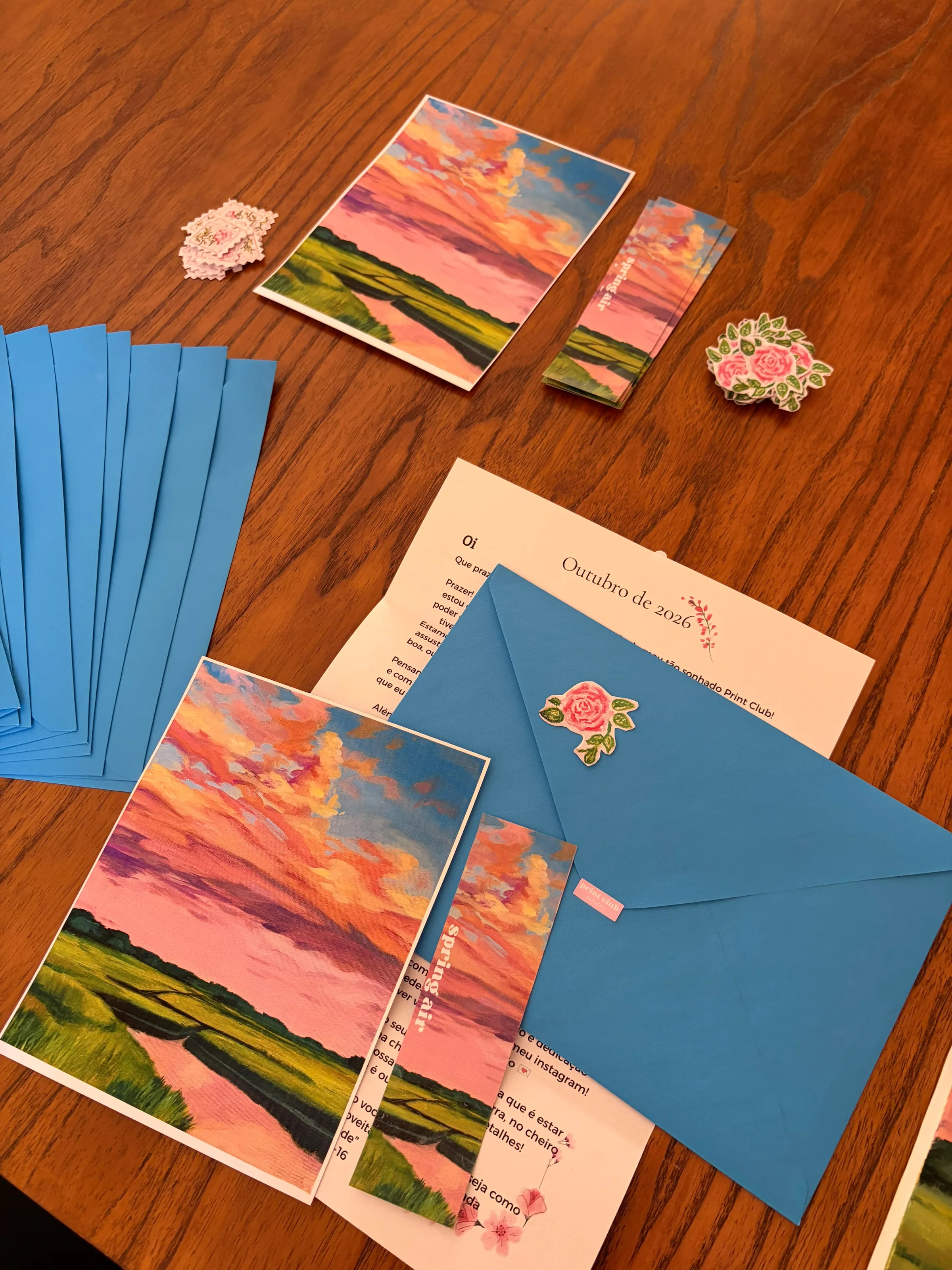Kit do Print Club de outubro sobre uma mesa de madeira, com prints, marcadores, adesivos de rosa, envelopes azuis e a carta.
